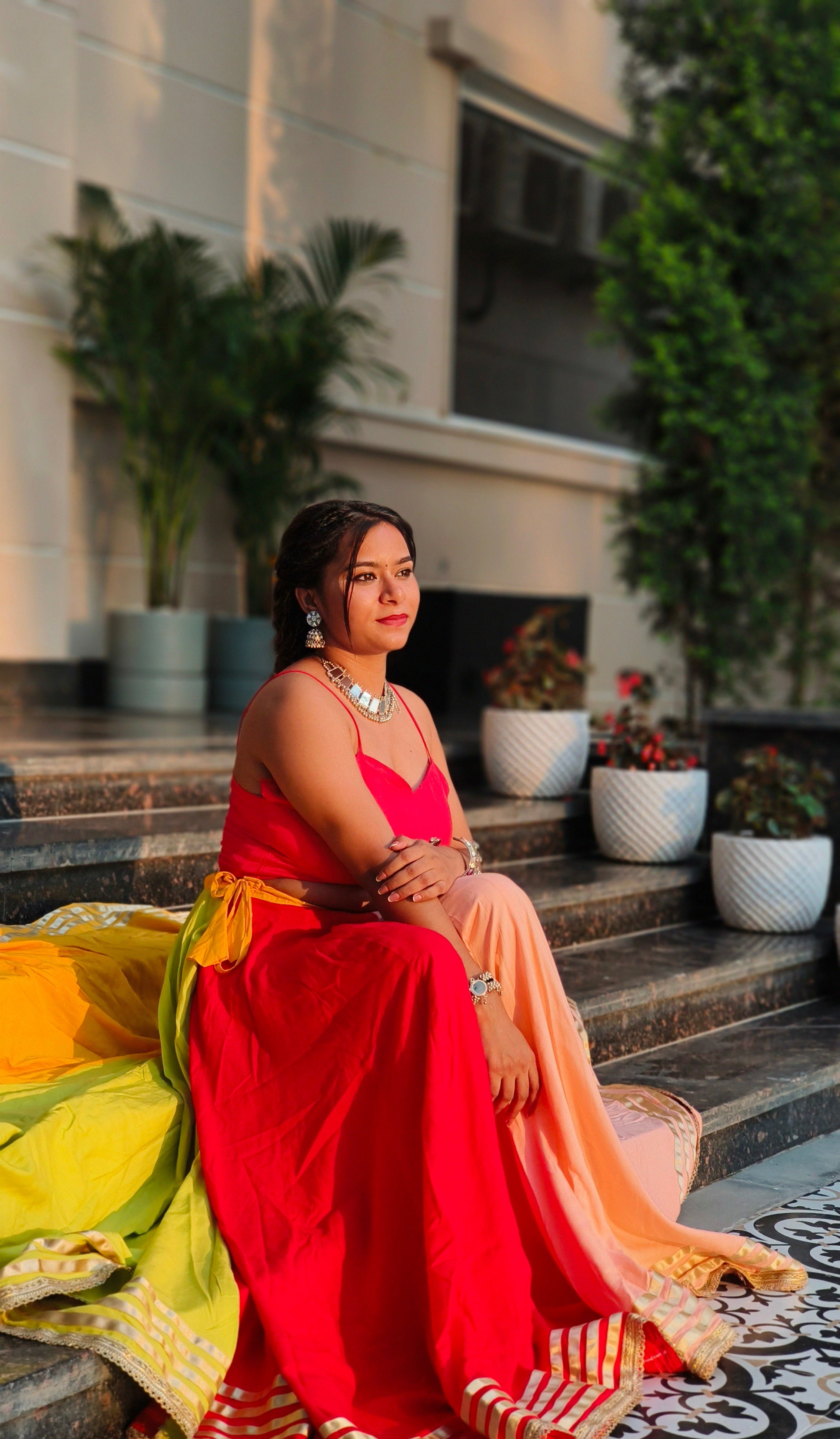 Woman in a red saree sitting on steps with plants and a building in the background